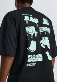 Svart t-shirt med mintgröna grafiska tryck av olika stolar och bänkar på ryggen. Texten lyder "THE SUNDAY CLUB" och "SERVING THE GAME FOR GENERATIONS."