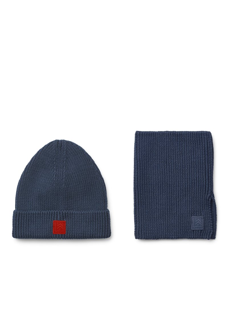 Πλεκτό ναυτικό σκουφί με διπλωμένο ρέλι και κόκκινο λογότυπο, συνδυασμένο με έναν αντίστοιχο ναυτικό ριγωτό σκ scarf που διαθέτει μια μικρή λεπτομέρεια λογότυπου.