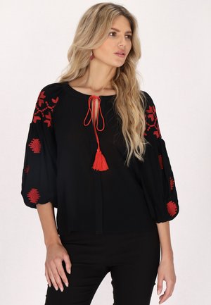 Schwarze Bluse mit roter Stickerei an den Ballonärmeln und einer Quastenbindedetail am Ausschnitt. Leichter Stoff und lässige Passform.