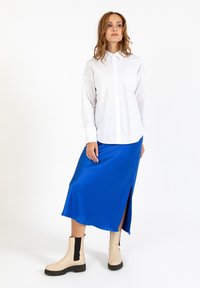 Witte button-up shirt gecombineerd met een blauwe satijnen midi rok, voorzien van een zijsplit, aangevuld met beige enkellaarzen met zwarte accenten.