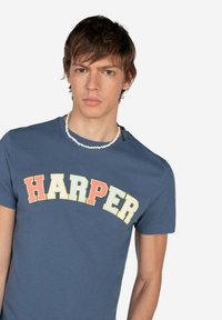 Camiseta de algodón azul con un logo curvado de "HARPER" en tonos rojo, amarillo y amarillo. Manga corta y escote redondo.
