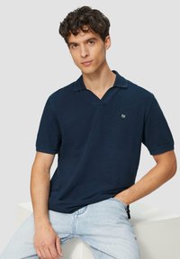 Tumedat sinised lühikeste varrukatega polo-särgid V-kaelusega, tekstuurne kangas, peene logodetailiga ning lõdva lõikega, koos heledate siniste teksadega.