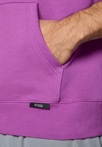 Sweatshirt roxo com bolso frontal, bainha ribbed e costuras visíveis. Apresenta uma etiqueta "CITY EDITION" perto da parte inferior. Textura suave.