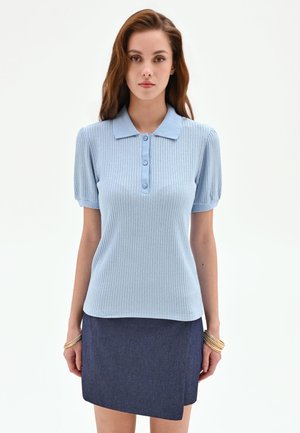 Donna che indossa una polo a maniche corte azzurro chiaro a coste e una gonna asimmetrica blu scuro, in piedi davanti a uno sfondo bianco semplice.