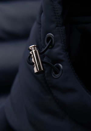 Dettaglio di un giubbotto imbottito blu navy con un fermacorda metallico, cordino elastico nero e asola, che mette in mostra un tessuto liscio e testurizzato.