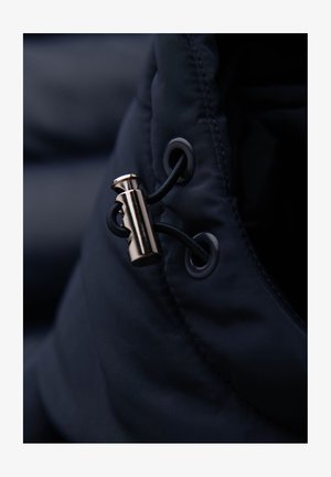 Dettaglio di un giubbotto imbottito blu navy con un fermacorda metallico, cordino elastico nero e asola, che mette in mostra un tessuto liscio e testurizzato.