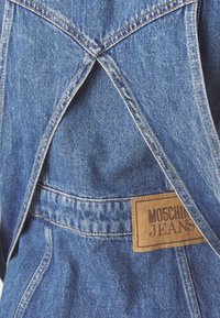 Džinsu kombinezons ir ar krustenisku muguras dizainu, gaiši zila auduma, oranžiem diegiem un brūnu ādas etiķeti ar uzrakstu "MOSCHINO JEANS."
