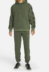Olivgrüner, flauschiger Kapuzenpullover mit Kängurutasche und schwarzen Akzenten an den Ärmeln, kombiniert mit passenden Jogginghosen und schwarzen Turnschuhen.