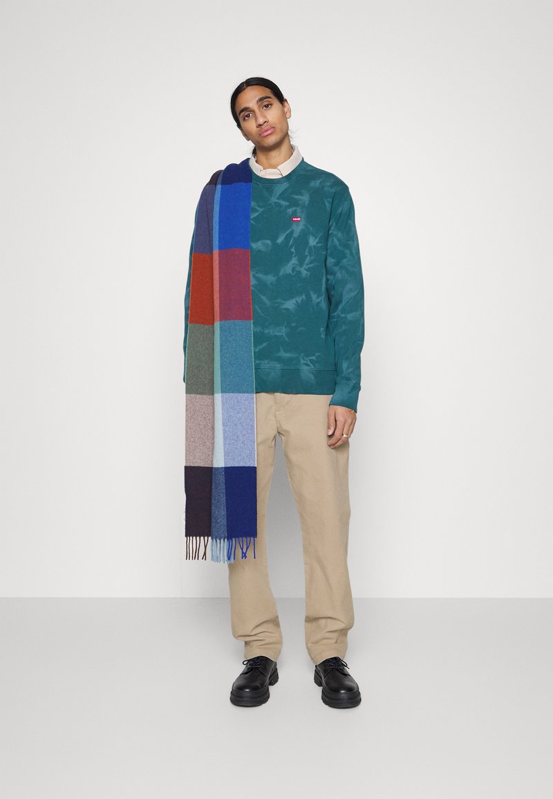 Sweatshirt turquoise à col rond avec un petit logo, pantalons beiges et une écharpe en laine multicolore avec des blocs de bleu, rouge et vert. Chaussures noires.