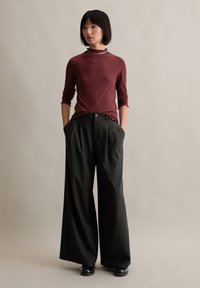 Bordeauxfarbenes, geripptes Langarmoberteil mit rüschigem Ausschnitt, kombiniert mit locker sitzenden, dunkelgrauen Hosen, die über Knopf- und Faltendetails verfügen.