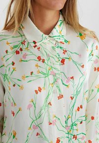 Chemise blanche à boutons avec des tiges vertes et de petites imprimés de fleurs rouges, jaunes et roses portée par une personne aux longs cheveux blonds.