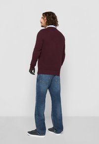 Burgundi pulóver texturált kötésével, kerek nyakkal, kék farmernadrággal párosítva. A modellen fekete kesztyű és sötétkék cipő van.