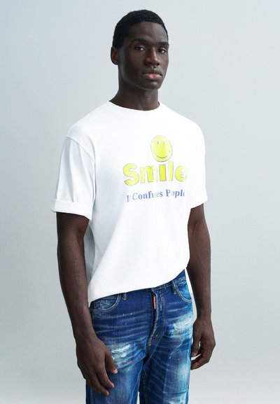 T-shirt blanc à manches courtes avec un graphisme de visage souriant jaune et un texte bleu. Porté avec un jean bleu déchiré affichant différentes textures.