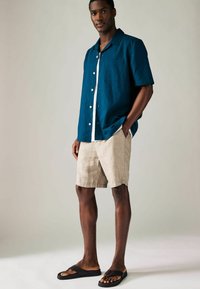Köksblå kortärmad skjorta med knäppning och ficka, beiga linne-shorts och svarta flip-flops. Slät textur och avslappnad passform.