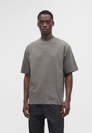 Ung mand iført en ensfarvet grå oversized t-shirt med korte ærmer og mørke bukser, stående foran en ensfarvet lys baggrund.