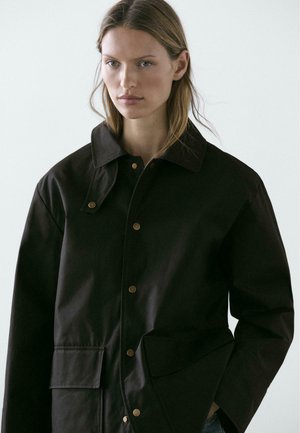 Junge Frau trägt eine dunkel übergroße Jacke mit Druckknöpfen und Klappentaschen, steht vor einem schlichten, hellen Hintergrund.