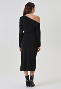 Schwarzes Ein-Schulter-Kleid mit langen Ärmeln, einem drapierten Rücken und einer taillierten Passform. Kombiniert mit schwarzen Stiefeletten mit Nietenakzenten.