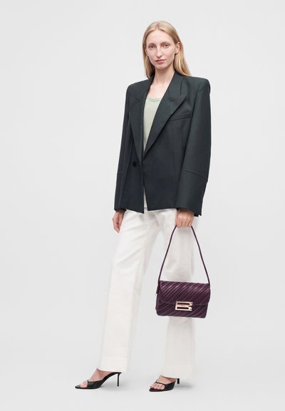 Γυναίκα με μαύρο oversized σακάκι, ανοιχτόχρωμο τοπ, άσπρο παντελόνι, μαύρα σανδάλια με τακούνι, κρατώντας μια μωβ τσάντα με χρυσό κούμπωμα σε απλό φόντο.