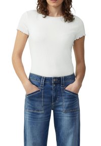 Frau mit lockigem Haar, die ein weißes, kurzärmeliges, geripptes Oberteil und blaue hochtaillierte Jeans mit großen Fronttaschen trägt, die Hände in den Taschen.