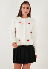 Witte gebreide cardigan met ronde hals, knopen aan de voorkant en rode kers-pompomversieringen. Gecombineerd met een zwarte plissé rok.