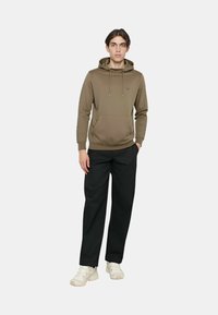 Felpa con cappuccio verde oliva, realizzata in tessuto liscio, con tasca a marsupio e coulisse. Abbinata a pantaloni neri e scarpe chiare.