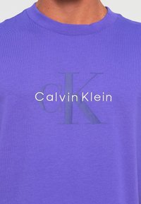 Krupni plan ljubičaste majice s okruglim izrezom s blagim reljefnim "CK" logom i bijelim "Calvin Klein" tekstom koji je centriran na prsima.