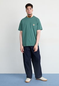 Green cotton T-shirt with a chest pocket, paired with dark blue denim pants. White sneakers complete the outfit. Simple, relaxed fit design.