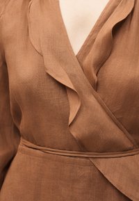 Καφέ φόρεμα τύπου wrap με βολάν στη V-λαιμόκοψη και ζώνη από το ίδιο ύφασμα στη μέση, κατασκευασμένο από υφή λινό υλικό.