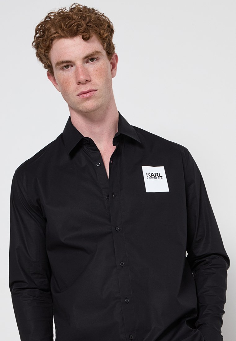 Chemise noire à manches longues en tissu lisse. Présente un design à boutons et un logo blanc brodé sur la poitrine gauche.