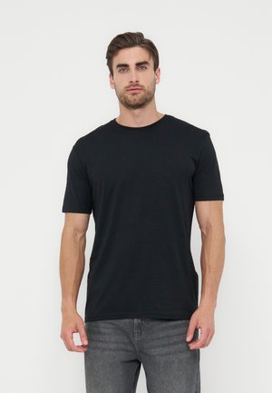 Mand med kort brunt hår iført en ensfarvet sort kortærmet T-shirt og grå jeans, stående mod en hvid baggrund.