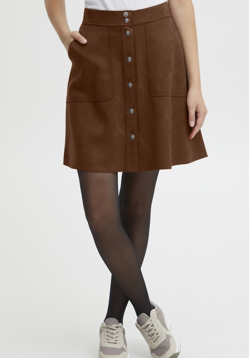 Brown suede skirt with a flared shape, front buttons, and two side pockets. Paired with black tights and light-colored sneakers.
