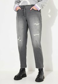 Jean en denim gris avec une coupe fuselée, présentant des déchirures et une taille haute. Associé à des bottines noires et un pull gris à motifs.