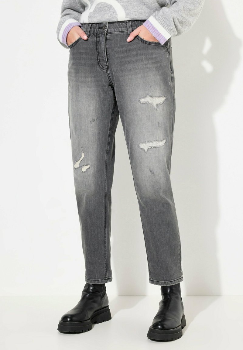Jean en denim gris avec une coupe fuselée, présentant des déchirures et une taille haute. Associé à des bottines noires et un pull gris à motifs.