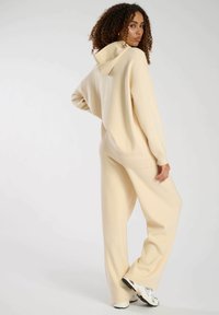 Sudadera de color crema, de corte oversize, con capucha y puños acanalados, combinada con pantalones de pierna ancha a juego, que presentan una textura suave y un ajuste relajado.