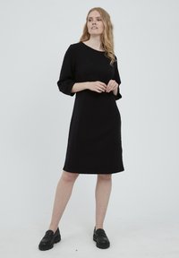 Musta polvipituinen mekko, jossa on teksturoitu kuosi, leveät kolmannespuolihihat ja pyöreä kaula-aukko, yhdistettynä mustiin paksupohjaisiin loafer-kenkiin.