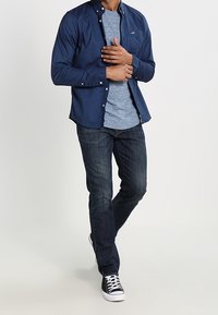 Man i marinblå knapp-up skjorta över en blå t-shirt, mörka jeans och svarta sneakers, står med händerna knäppta nära bröstet.