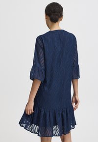 Rochie albastră închis cu un design fluid, având un model texturat cu frunze, mâneci trei sferturi și un tiv volanat. Material semitransparent.