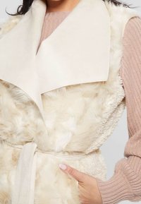 Donna che indossa un gilet in pelliccia sintetica beige sopra un maglione a coste rosa chiaro, con una mano in tasca, colletto ampio e cintura in tessuto annodata in vita.