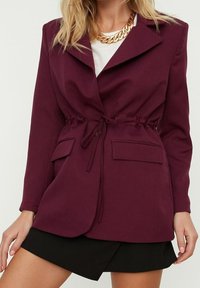 Blazer bordeaux avec une taille ajustable par cordon de serrage, deux poches avant et un col à revers, porté sur une chemise blanche et une jupe noire.