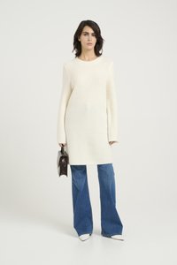 Pulover crem cu mâneci lungi, cu un design tricotat texturat, asortat cu blugi cu croială evazată de culoare albastră și pantofi albi cu vârful ascuțit, ținând o geantă de mână neagră.