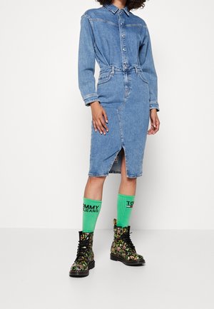 Vrouw die een blauw denimjurk draagt met knopen en een ceintuur in de taille, neon groene Tommy Jeans sokken en zwarte, bloemetjesmotief veterlaarzen, staand tegen een witte achtergrond.