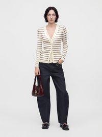 Femme portant un cardigan crème à rayures, un jean large foncé, des mocassins noirs, tenant un petit sac à main rouge foncé sur un fond clair uni.