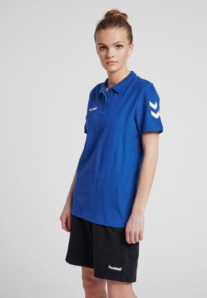 Persona joven de pie con camiseta polo azul y pantalones cortos negros con logotipos de la marca sobre un fondo claro y liso.