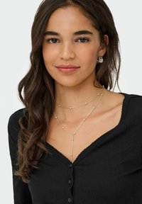 Srebrny naszyjnik z warstwowych okrągłych koralików, noszony z czarnym prążkowanym topem z falistym dekoltem i guzikami. Włosy lekko falowane.