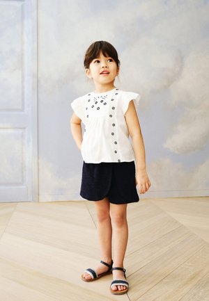 Camicia bianca a maniche corte con ricami floreali, abbinata a pantaloni corti blu scuro e sandali neri con dettaglio intrecciato e fascia.