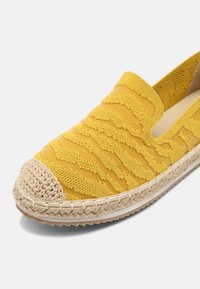 Geltoni slip-on batai su tekstūruotu audiniu ir pintu jutos viduriu. Apvalus pirštų dangtelis su nėrinių akcentais.