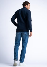 Navy ribbed pullover with a high collar, paired with light blue jeans and white sneakers, viewed from the back.