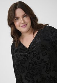 Fekete felső virágmintával, texturált anyagból. V-alakú nyakkivágása van, ráncolt részletekkel és hosszú ujjakkal.