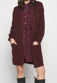 Bordowy sweter z dzianiny z otwartym przodem, długimi rękawami i bocznymi kieszeniami, noszony na koszulowej sukience w pasującym kolorze.