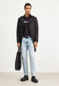 Abrigo negro sobre una camiseta negra, jeans de un azul claro y botas negras de plataforma. Sostiene una bolsa tote negra. Diseño y colores simples y modernos.